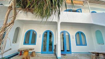 A building with white walls and blue accents, featuring arched windows and doors, a stone courtyard, and rustic wooden furniture.