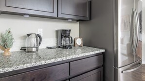A kitchen with dark cabinets, a stainless steel refrigerator, a coffee maker, and a kettle on a granite countertop. - Cozy Modern Home! (Vancouver)