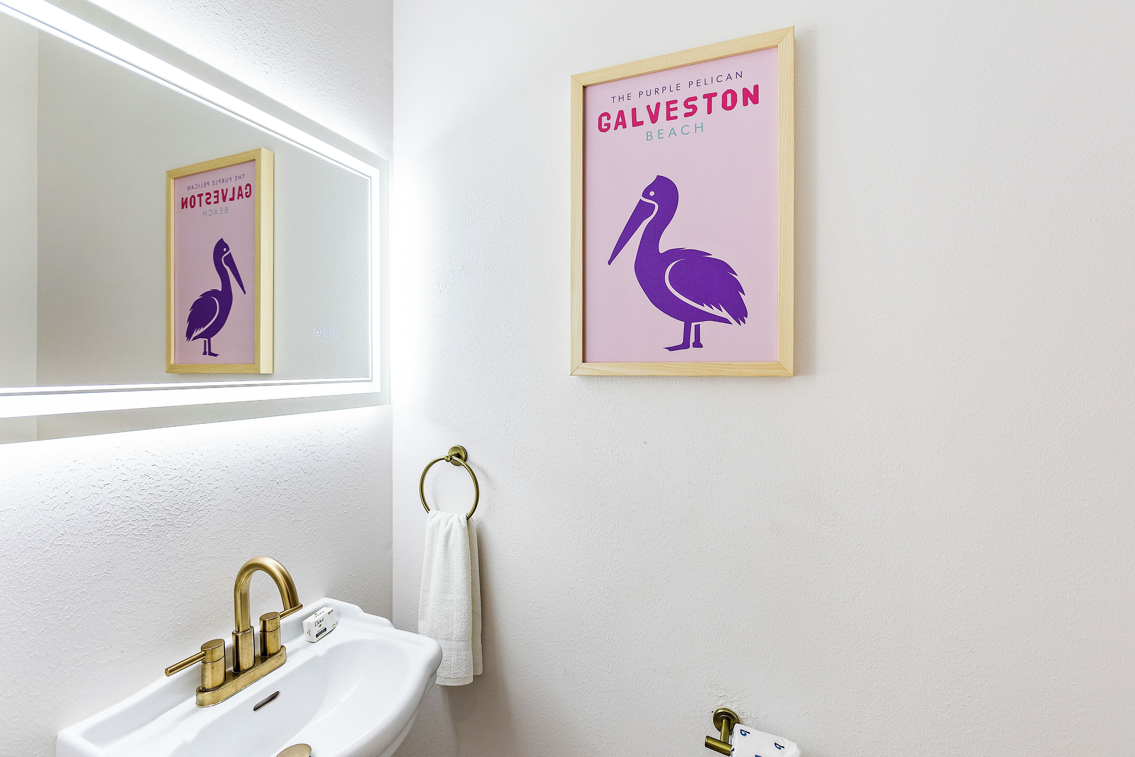 A bathroom with a white sink, gold faucets, and a framed poster of a pelican on the wall.