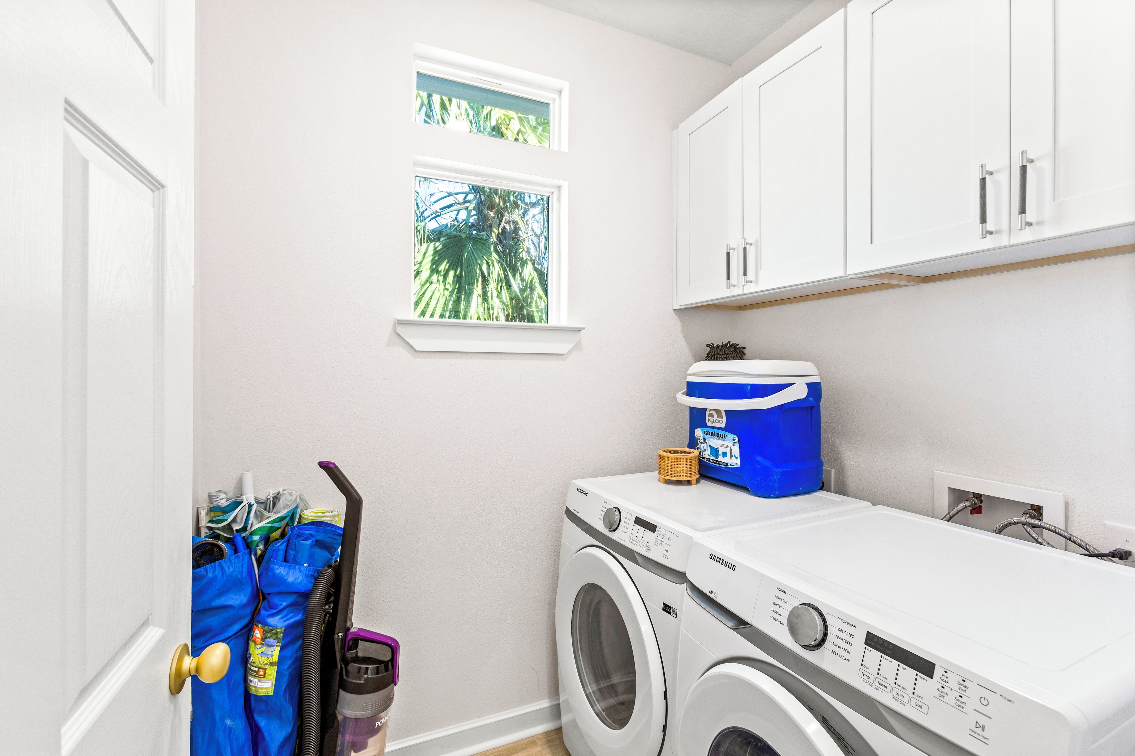 A laundry room with a Samsung washing machine, a blue container, and a window with a view of palm trees.