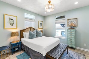 A bedroom with a bed, bedside table, lamp, dresser, and artwork on the walls. - The Purple Pelican (Galveston)