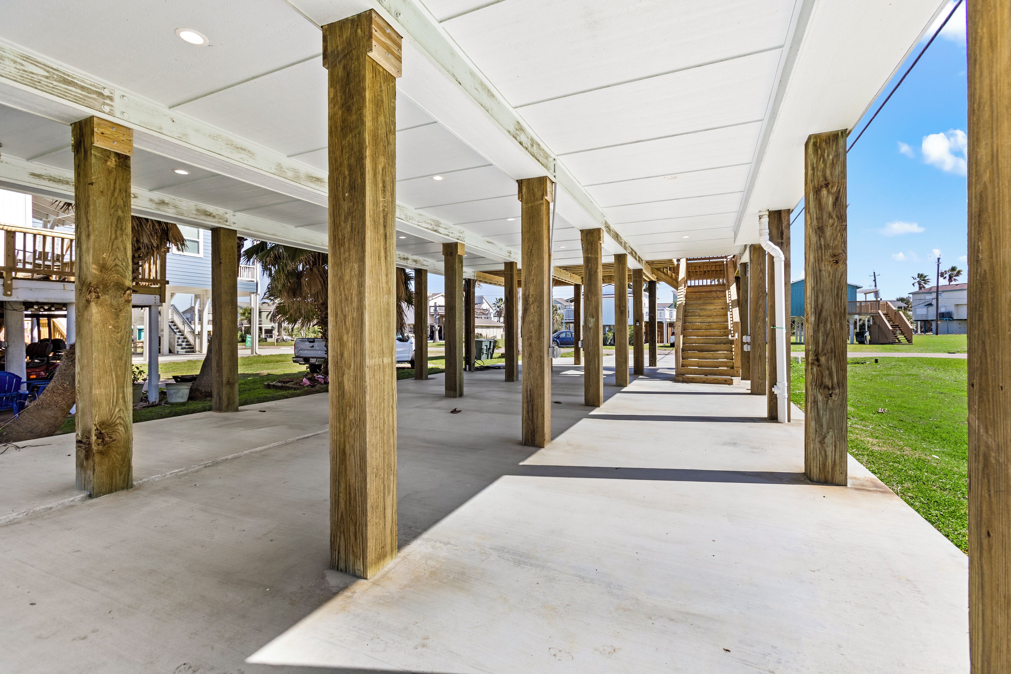 A covered walkway with wooden pillars, a concrete floor, and a view of the surrounding area including a building and a grassy field.