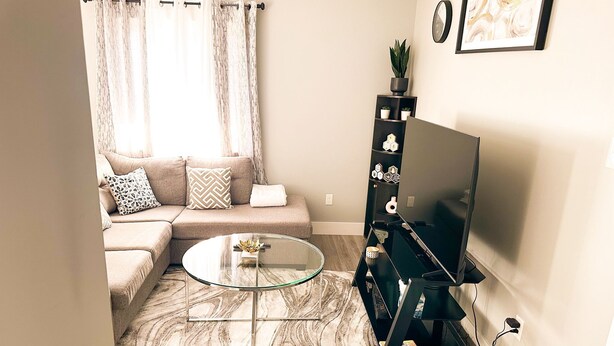 Een woonkamer met een beige bank, een glazen salontafel, een zwart tv-meubel en een vloerkleed met een marmerlook.