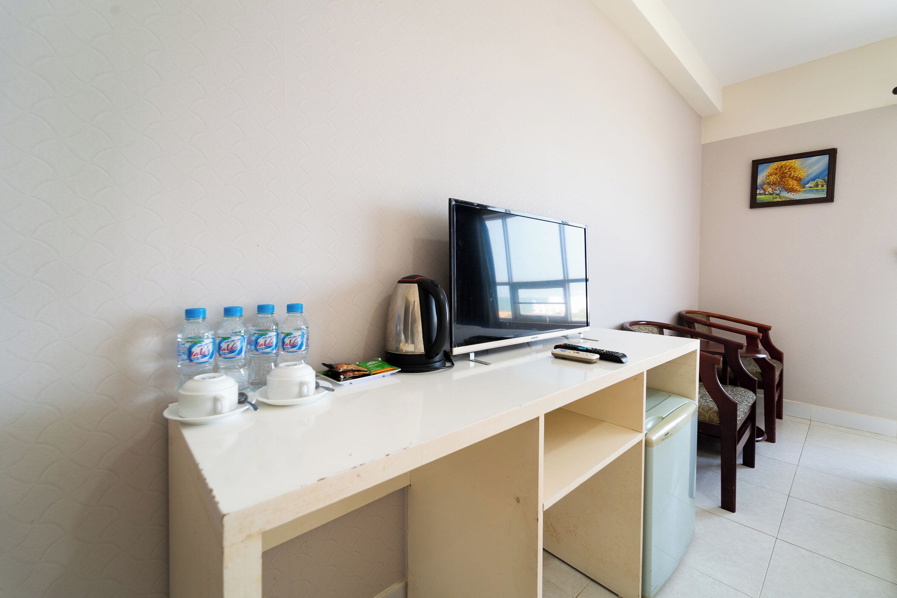 A room with a TV, water bottles, a kettle, a remote, and a painting on the wall.