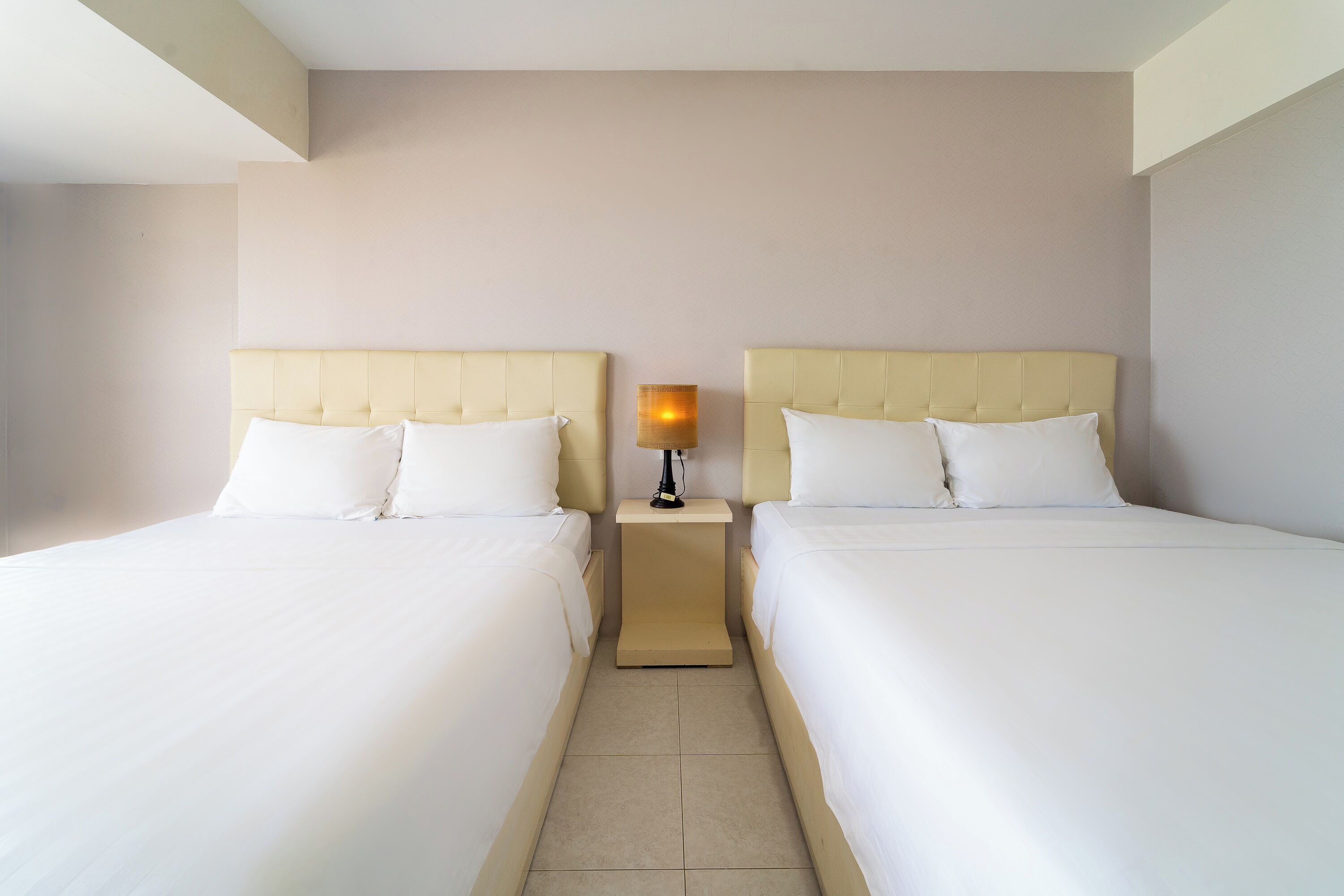 Habitación de hotel con dos camas, una mesita de noche con lámpara y una pared lisa.
