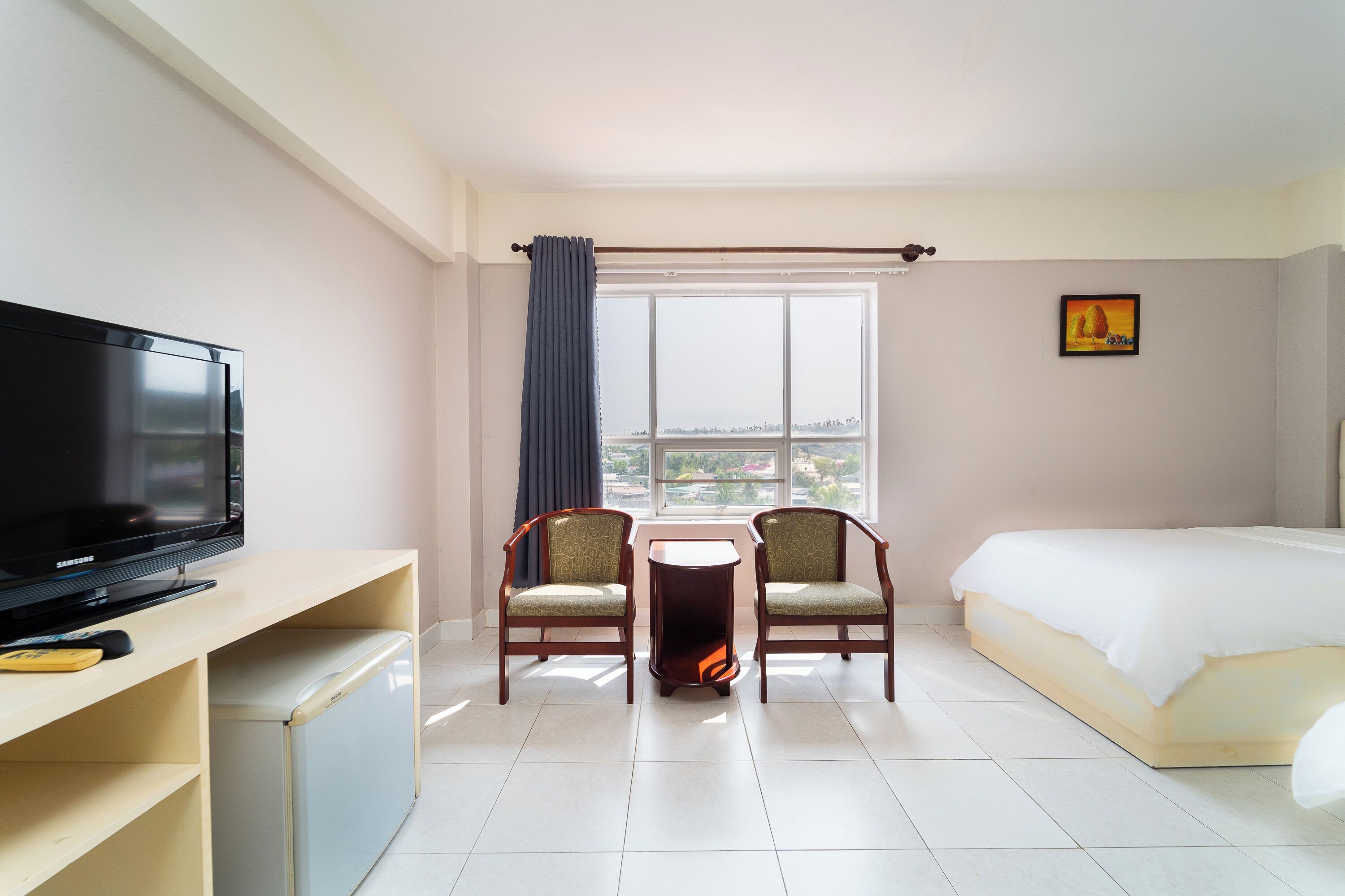 Une chambre d’hôtel avec un lit, une télévision, deux chaises, une petite table et une fenêtre avec des rideaux.