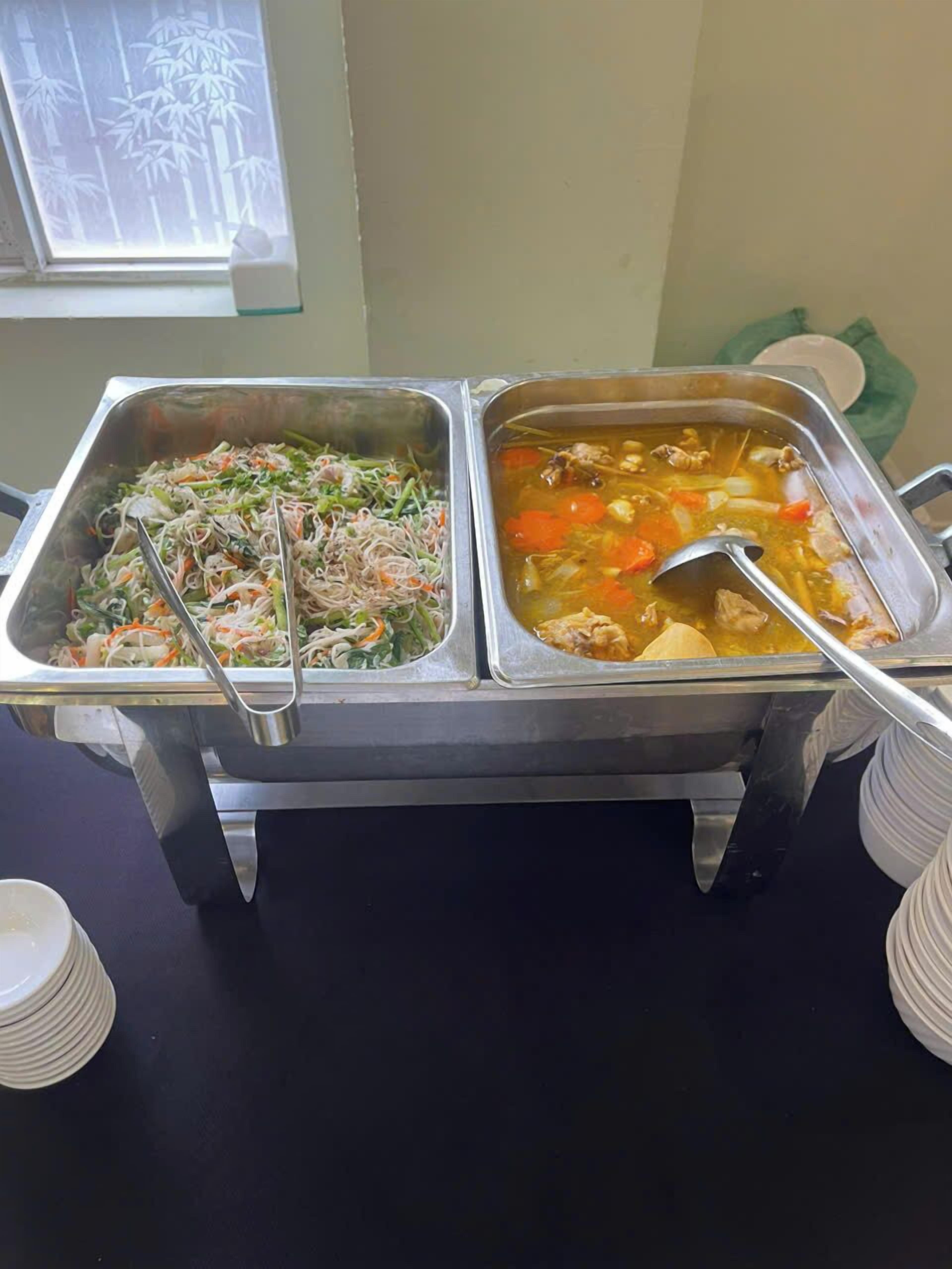Un bufé con dos bandejas de metal, una con un plato de fideos y la otra con sopa de verduras y carne.