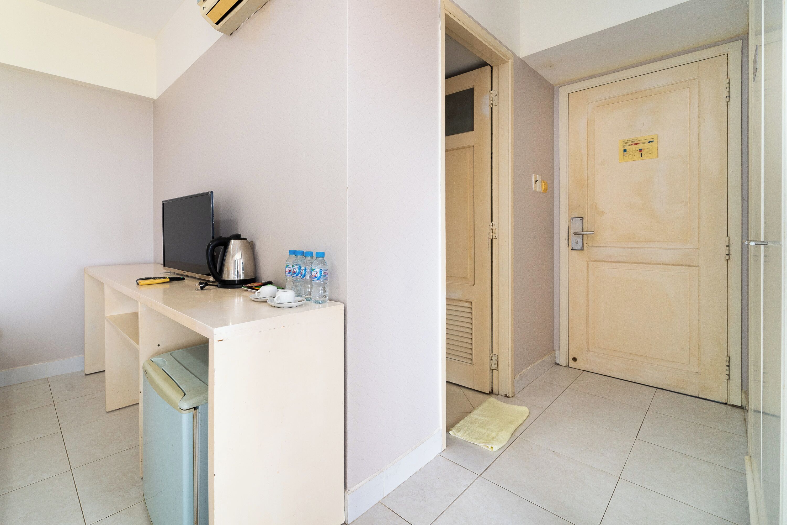 A small kitchenette with a coffee maker, water bottles, and a TV. There is a wooden door and a tiled floor.