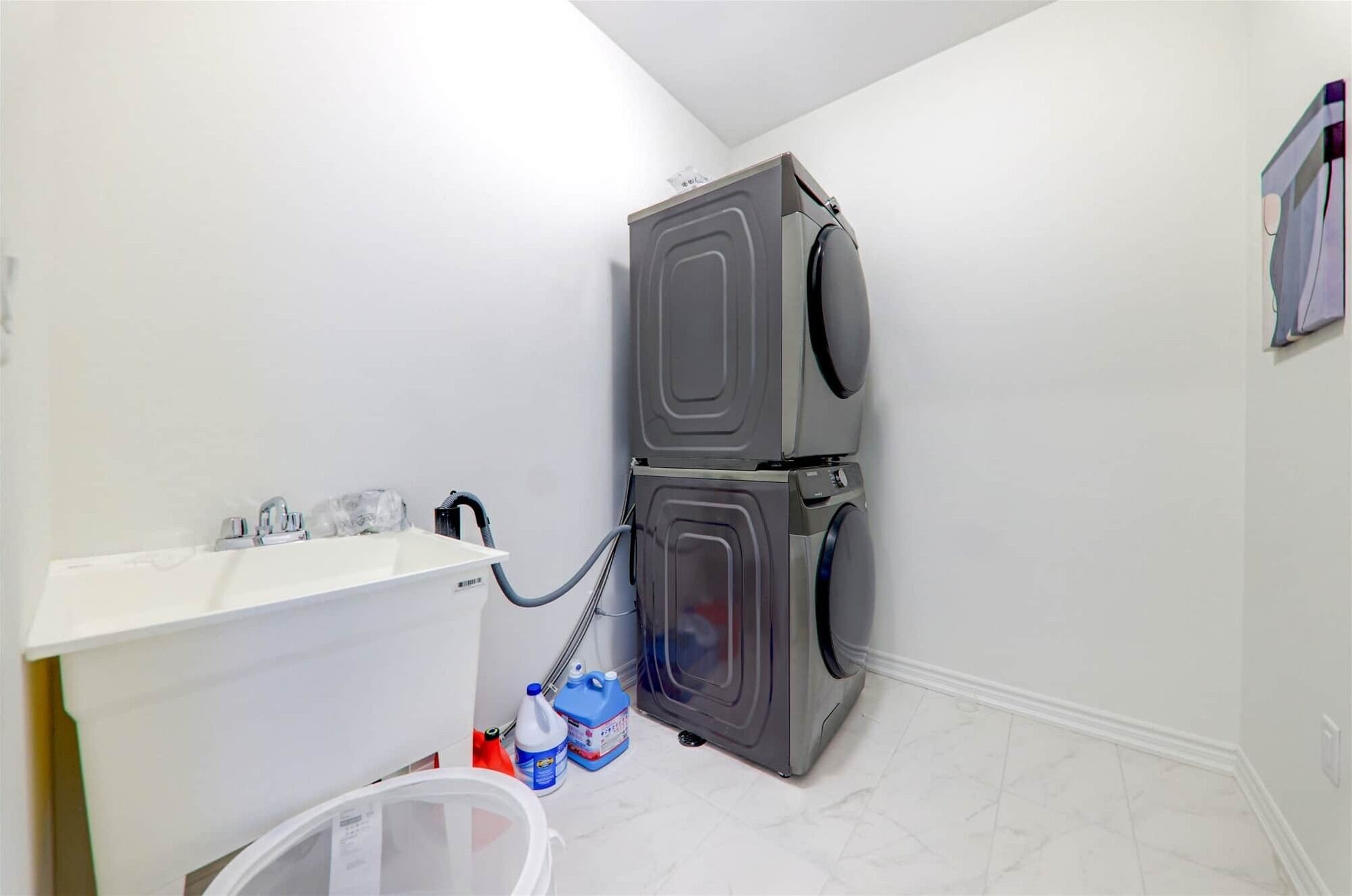 Un lavadero con dos lavadoras de carga frontal apiladas, un lavamanos blanco con grifo y un cubo de basura.