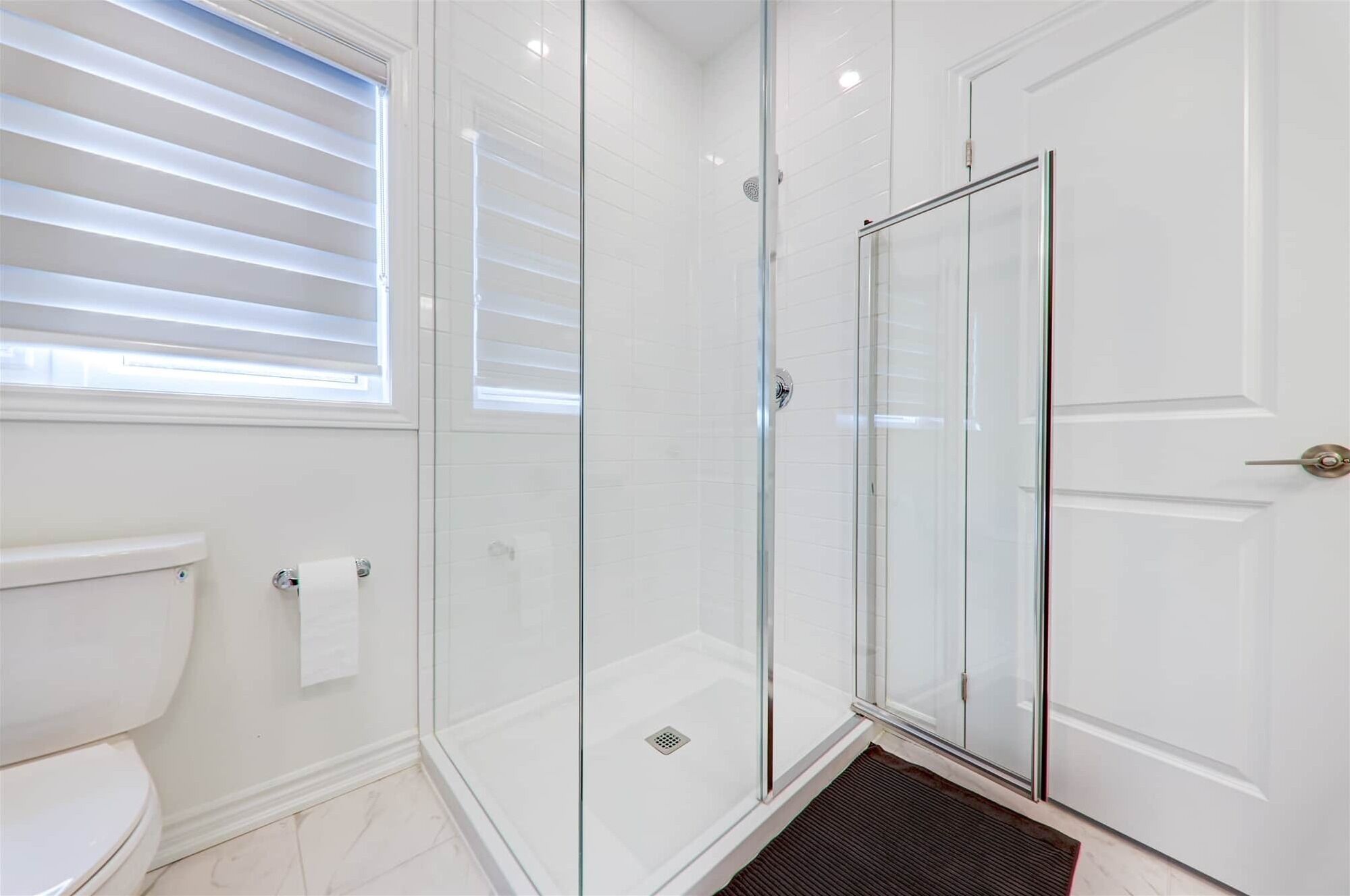 Een moderne badkamer met een douchecabine van glas, witte betegelde wanden, een toilet en een raam met zonwering.