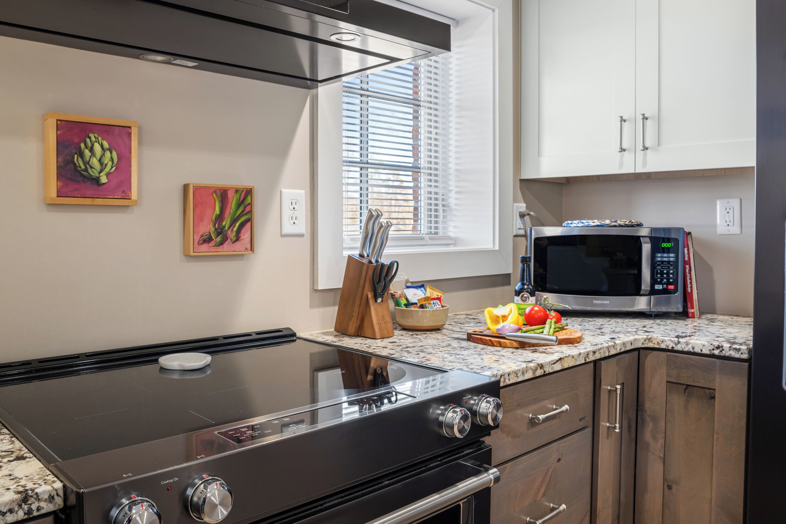 A kitchen with a stove, microwave, and cabinets. There are two paintings on the wall and a window with blinds.
