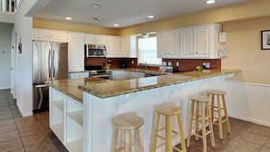 A spacious kitchen with white cabinets, a granite island with bar stools, stainless steel appliances, and a window providing natural light. - Under the Sea (Galveston)