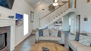 A spacious living room with a fireplace, a round wooden coffee table, a beige sectional sofa, and a staircase leading to an upper floor. - Under the Sea (Galveston)