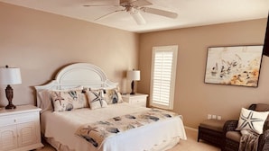 Une chambre à coucher avec un lit, des tables de chevet, une chaise, un ventilateur de plafond et un tableau accroché au mur.