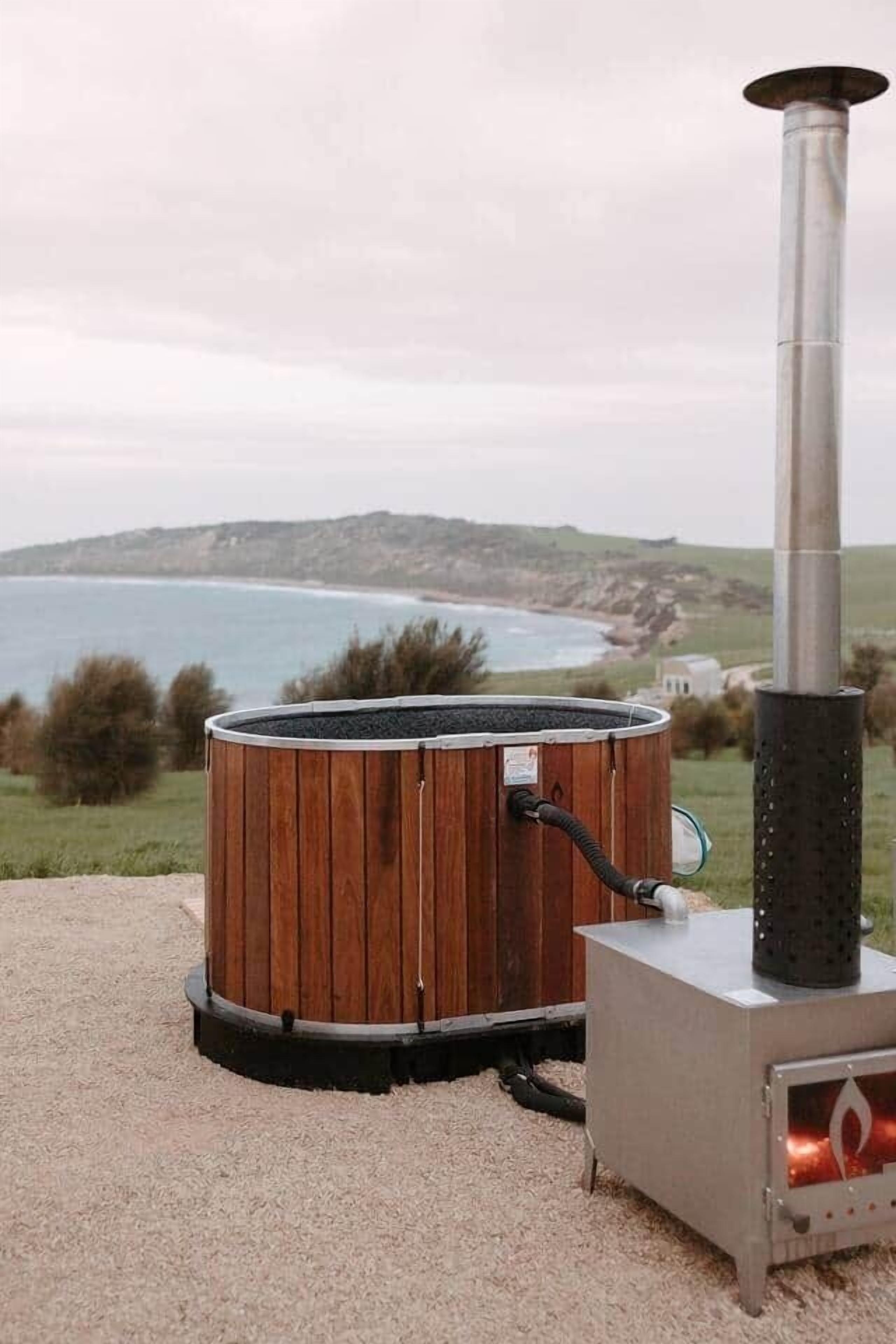 A wood-burning stove with a chimney, set on a gravel surface with a view of the sea and hills in the background.