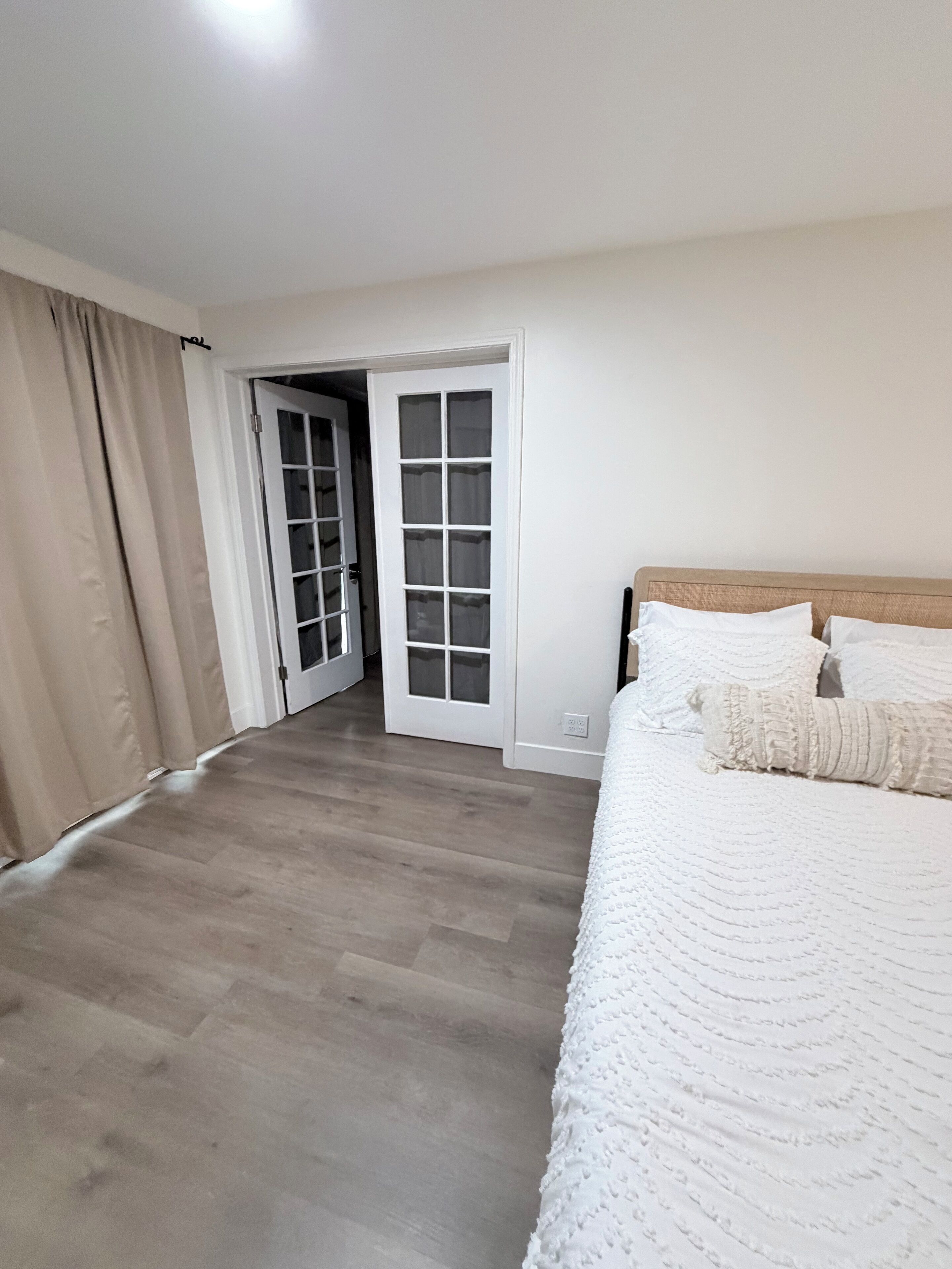 Un dormitorio con piso de madera, una cama con ropa blanca y una puerta que da a otra habitación.