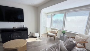 Un salon moderne avec un téléviseur à écran plat, une table basse ronde en bois, un canapé beige agrémenté de coussins et une grande fenêtre équipée de stores.