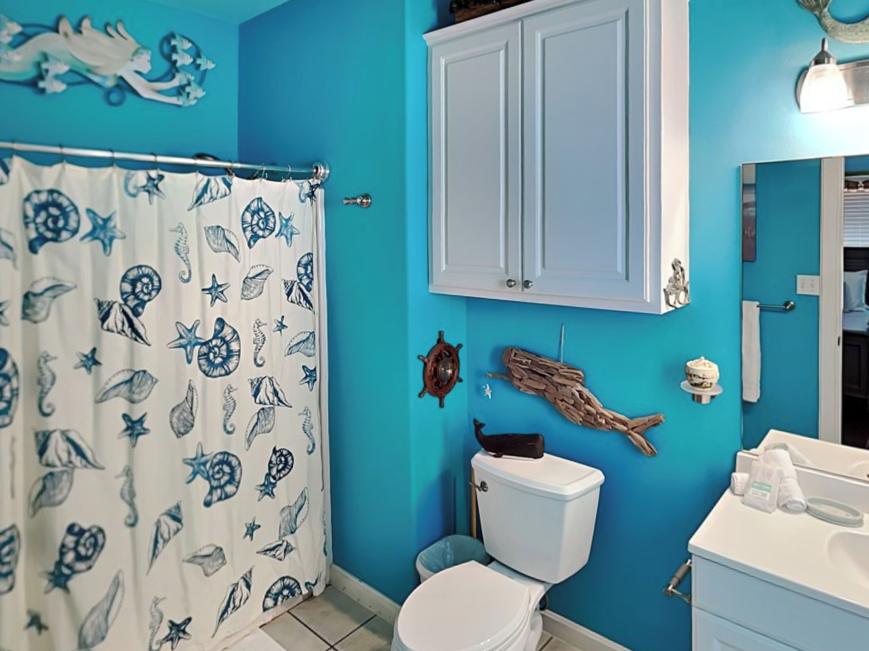 A bathroom with a blue wall, a white toilet, a shower curtain with a sea creature pattern, and a wooden fish decoration.