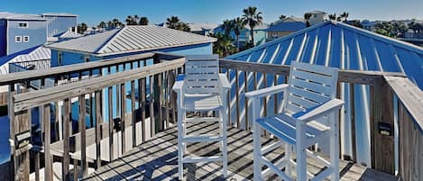 A wooden deck with white chairs, overlooking a coastal area with blue buildings and palm trees.