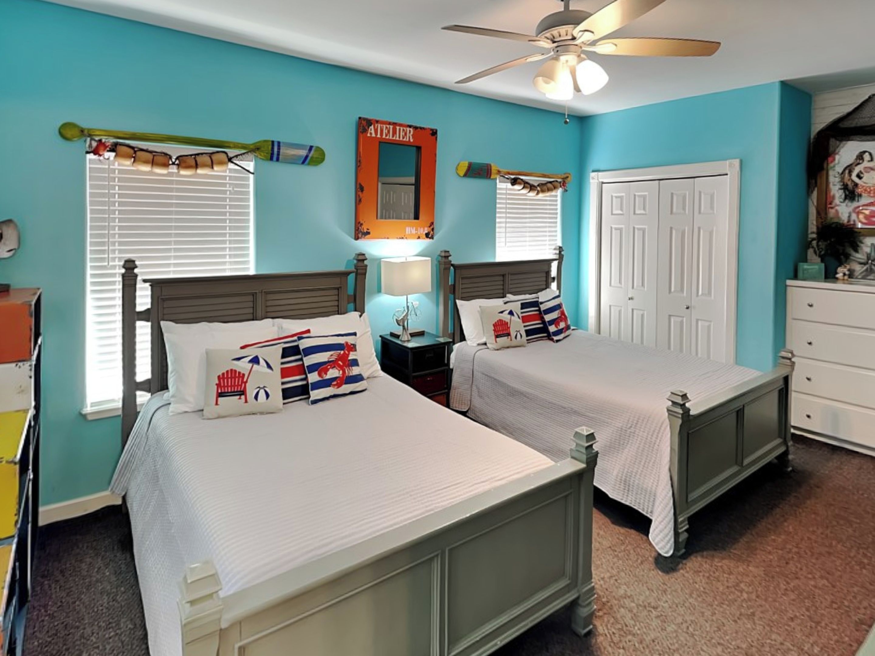 A bedroom with two beds, a ceiling fan, a window with blinds, a nightstand with a lamp, and a dresser.