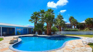 A swimming pool with stone tiles, palm trees, and a blue building in the background. - Coral Cabana (Port Aransas)