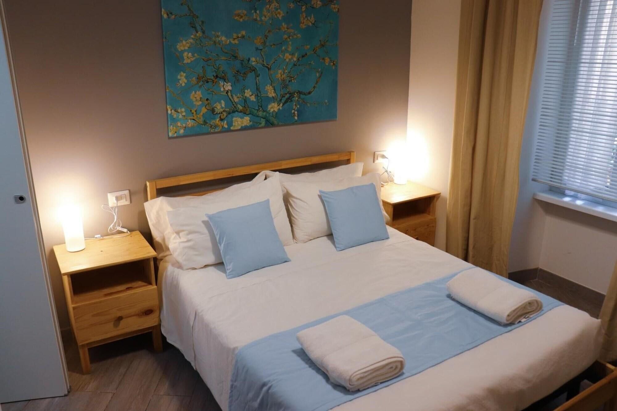 A neatly made bed with two pillows, a blue blanket, and two folded towels. There is a wooden nightstand with a lamp on each side of the bed. A painting hangs above the bed.