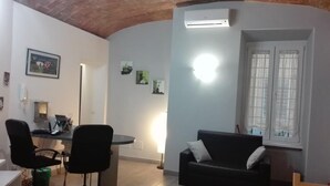 Un soggiorno con un divano in pelle nera, un tavolino rotondo e due sedie da ufficio nere. Si vede una finestra con tende bianche e un condizionatore d'aria fissato al muro.