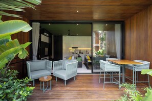 A modern living area with a wooden ceiling, a sliding glass door leading to a kitchen, and a seating arrangement with a sofa and two chairs. - Sunset Lagoon Rice Field Villa  (Ubud)