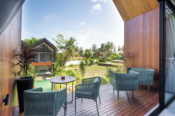 A wooden deck with wicker chairs and a table, overlooking a lush garden and a modern building. - Sunset Lagoon Rice Field Villa  (Ubud)