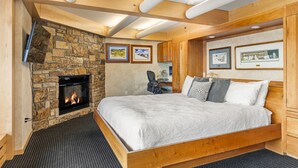 A bedroom with a stone fireplace, a large bed, wooden ceiling beams, and framed artwork on the walls. - Stonebridge Condos 1 Bdrm, Direct ski-in/ski-out (Snowmass Village)