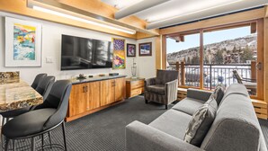 A modern living room with a large flat-screen TV, wooden cabinets, a gray sofa, and a dining area with a granite table. - Stonebridge Condos 1 Bdrm, Direct ski-in/ski-out (Snowmass Village)