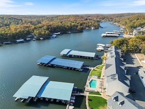 An aerial view of a lakeside property with docked boats, a swimming pool, and a large building with a sloped roof.
