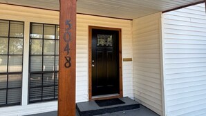 Una casa con puerta principal negra, un poste de madera con el número 5048 y una ventana con persianas.