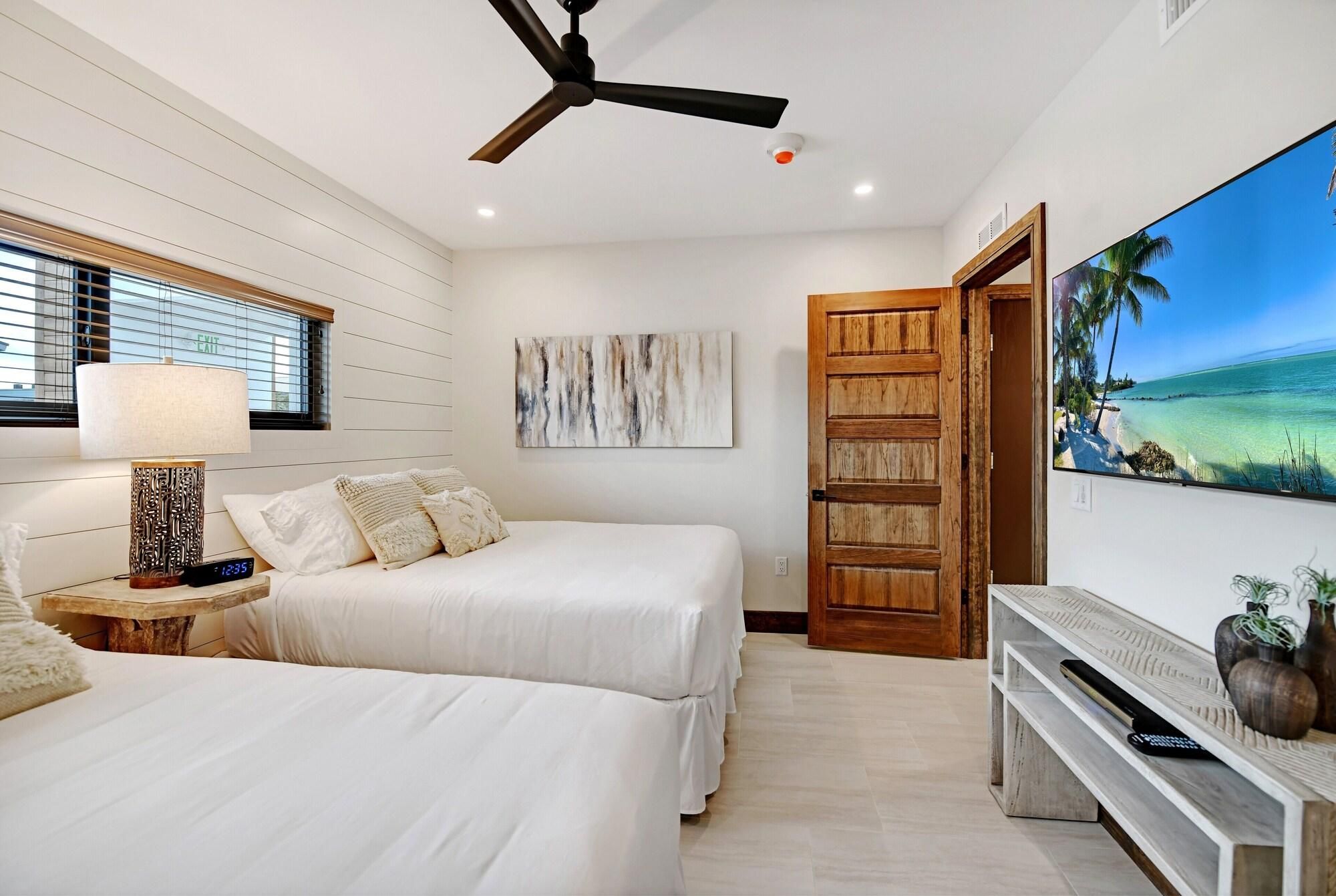 Une chambre d’hôtel avec deux lits, un ventilateur de plafond, un téléviseur fixé au mur qui diffuse une scène de plage, et une porte en bois.