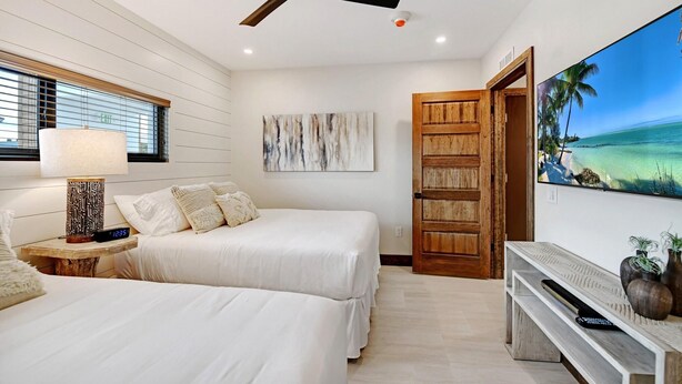 Une chambre d’hôtel avec deux lits, un ventilateur de plafond, un téléviseur fixé au mur qui diffuse une scène de plage, et une porte en bois.