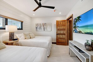 A hotel room with two beds, a ceiling fan, a wall-mounted TV displaying a beach scene, and a wooden door. - Bali Hai Beach Resort (Holmes Beach)