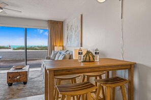 A modern room with a wooden dining table, two chairs, a small side table, and a view of the beach through large windows. - Bali Hai Beach Resort (Holmes Beach)