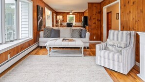 A cozy living room with a white sofa, a striped armchair, and a white bench. A kitchen is visible in the background with wooden walls and a ceiling fan. - 3 Mins to Watkins Glen | Seneca Wine Trail (Watkins Glen)