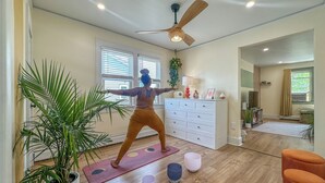 Un'ampia stanza con un ventilatore a soffitto, un'ampia pianta in vaso, un tappetino da yoga e una cassettiera bianca.