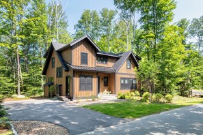 Une maison en bois à deux étages, dotée d’un toit en pente et entourée d’une zone boisée.