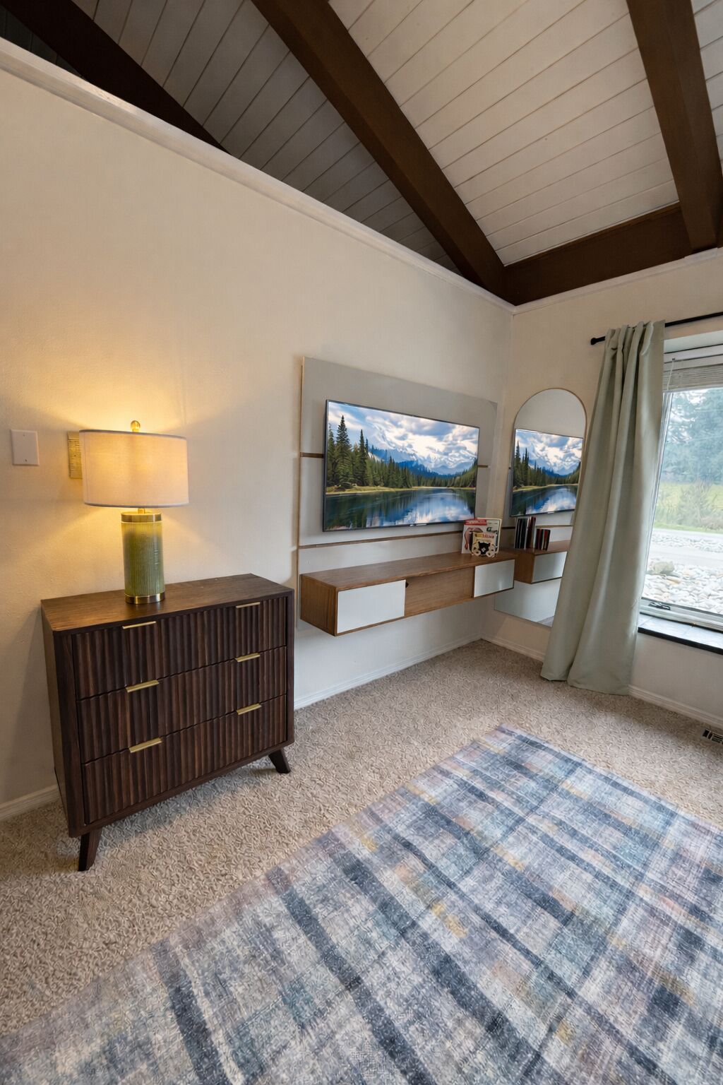 A room with a wooden dresser, a lamp, a painting, a mirror, and a window with curtains.