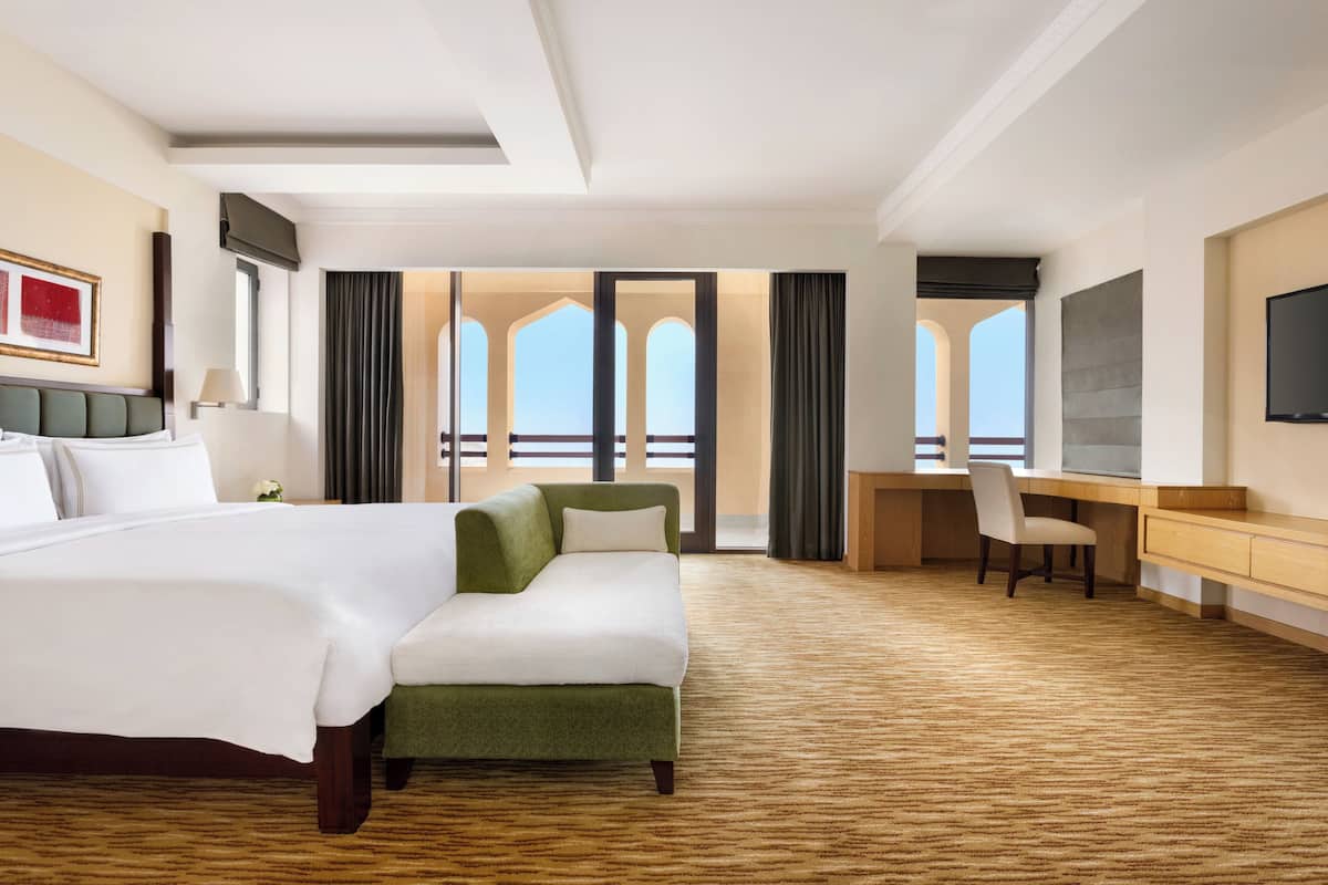 Habitación de hotel con una cama grande, un sofá verde, un escritorio y un televisor. Hay ventanas con cortinas y vistas al exterior.