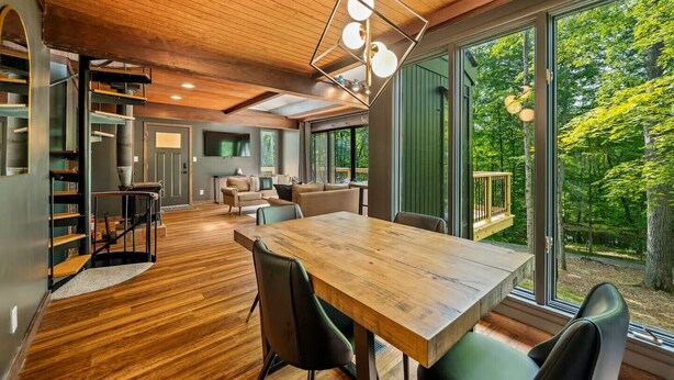 Amplia sala de estar con una mesa de comedor de madera, asientos cómodos y grandes ventanas que ofrecen vistas del bosque circundante.