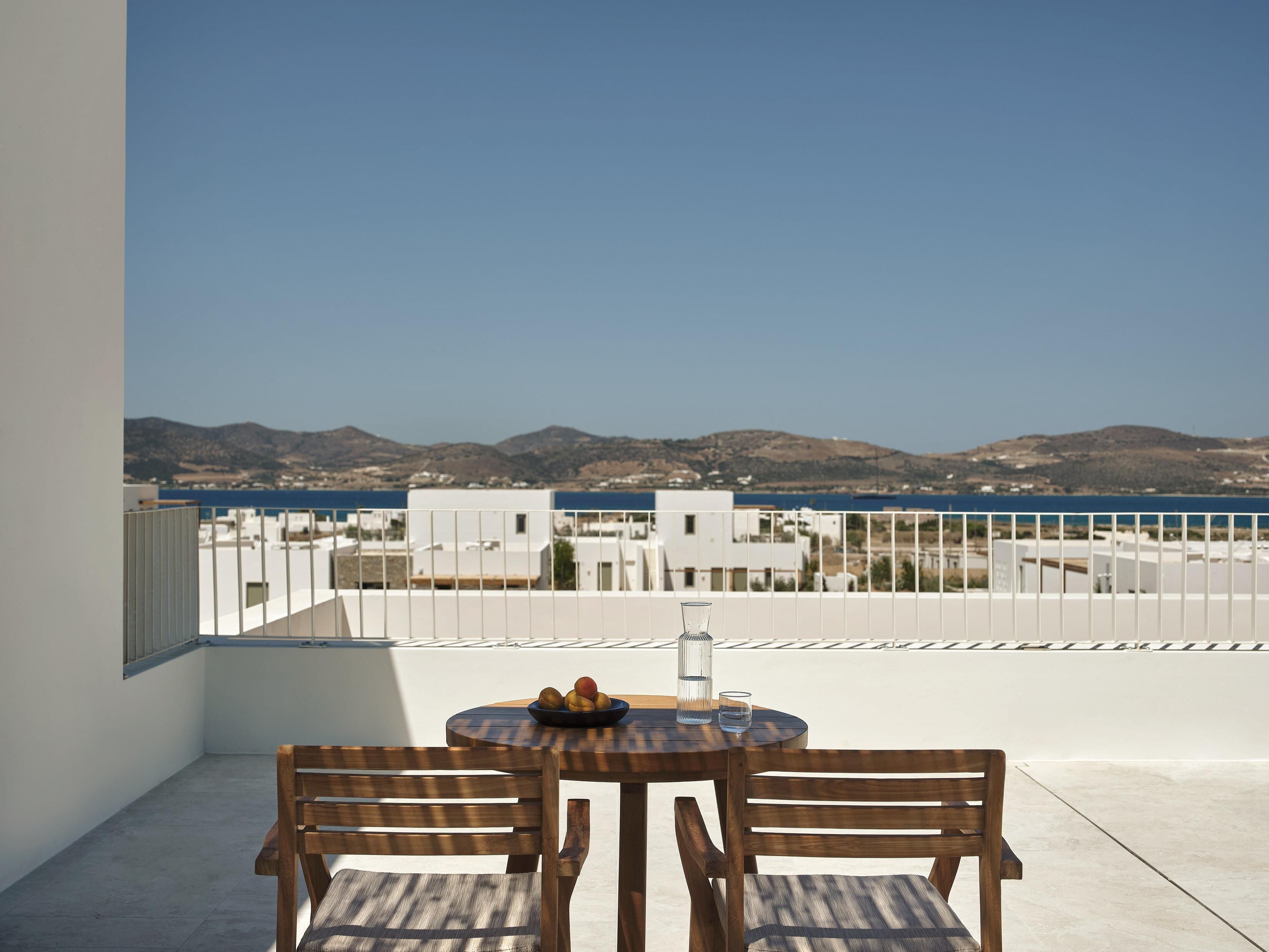 A balcony with two wooden chairs and a table, overlooking a coastal view with buildings and hills.