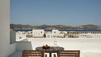 A balcony with two wooden chairs and a table, overlooking a coastal view with buildings and hills.