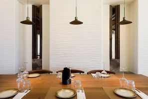 A dining table set for six with plates, cutlery, and glasses, in a room with white textured walls and hanging pendant lights.