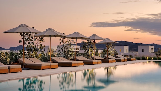 A poolside area with lounge chairs and umbrellas, a clear pool reflecting the surroundings, and a backdrop of mountains during sunset. - Parian Chronicle Hotel Paros, Destination by Hyatt (Paros)
