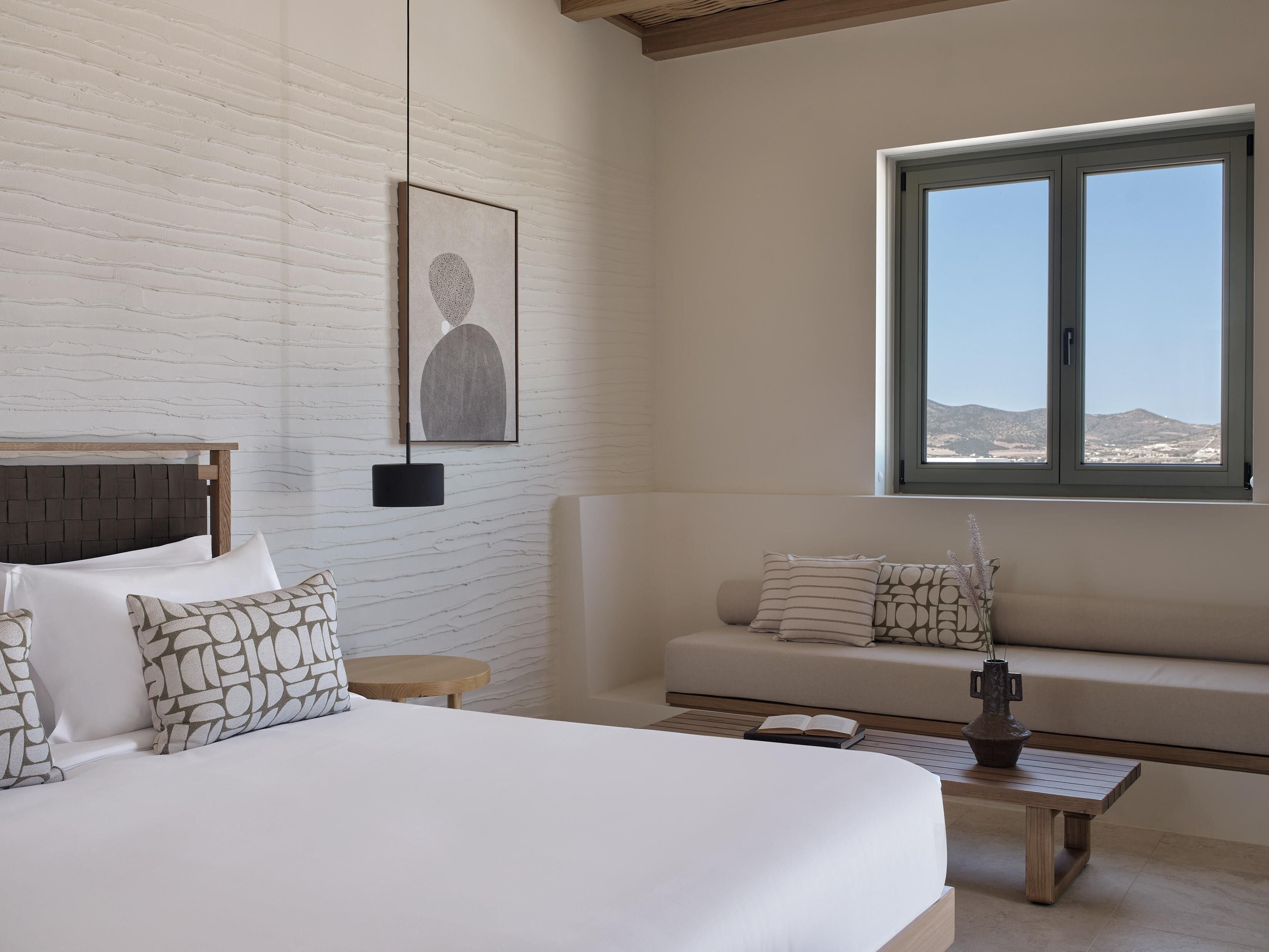 A bedroom with a large bed, a sofa, a small table, and a window with a view of the hills.