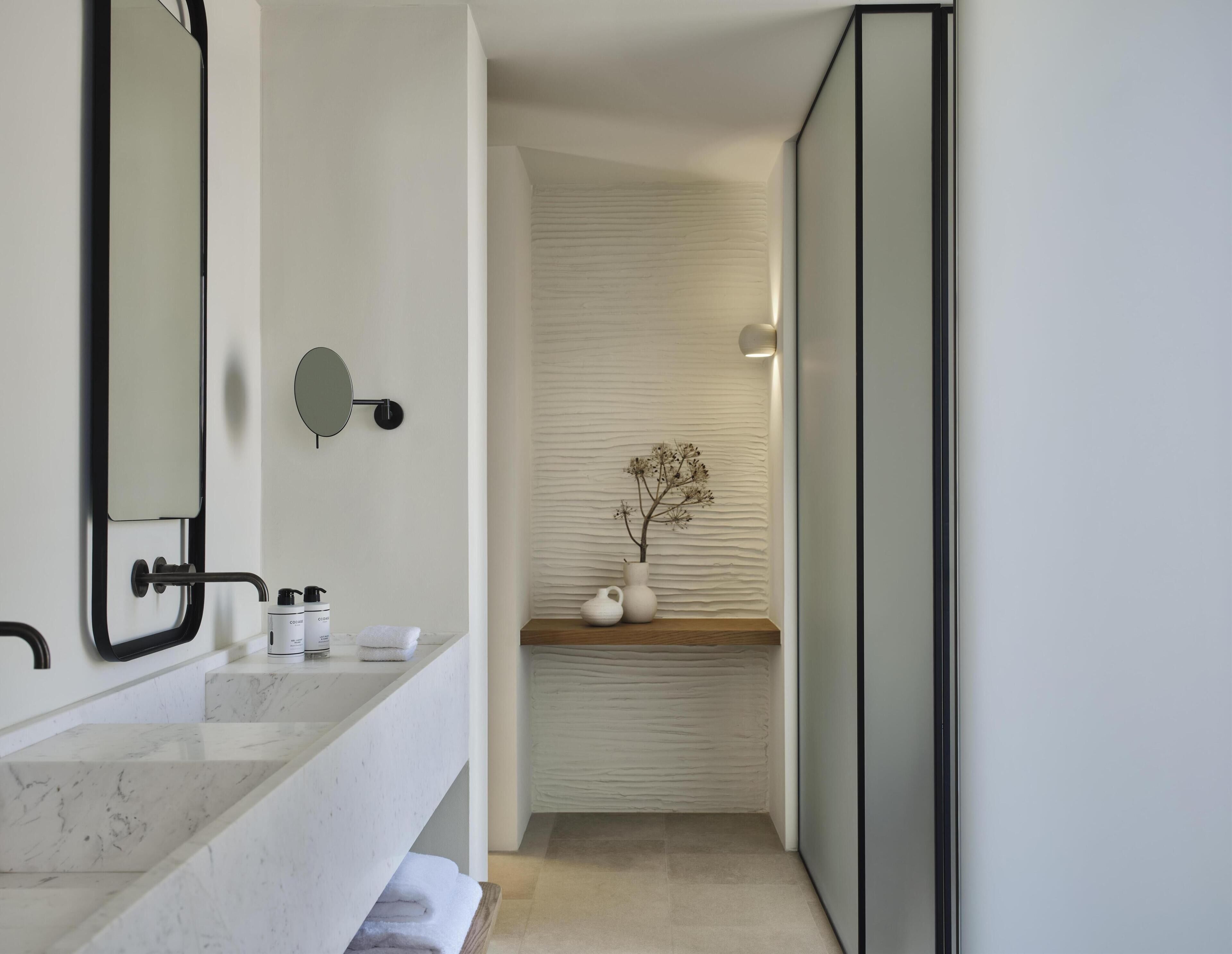 A modern bathroom with a large mirror, marble sink, and a vase with dried flowers on a shelf.