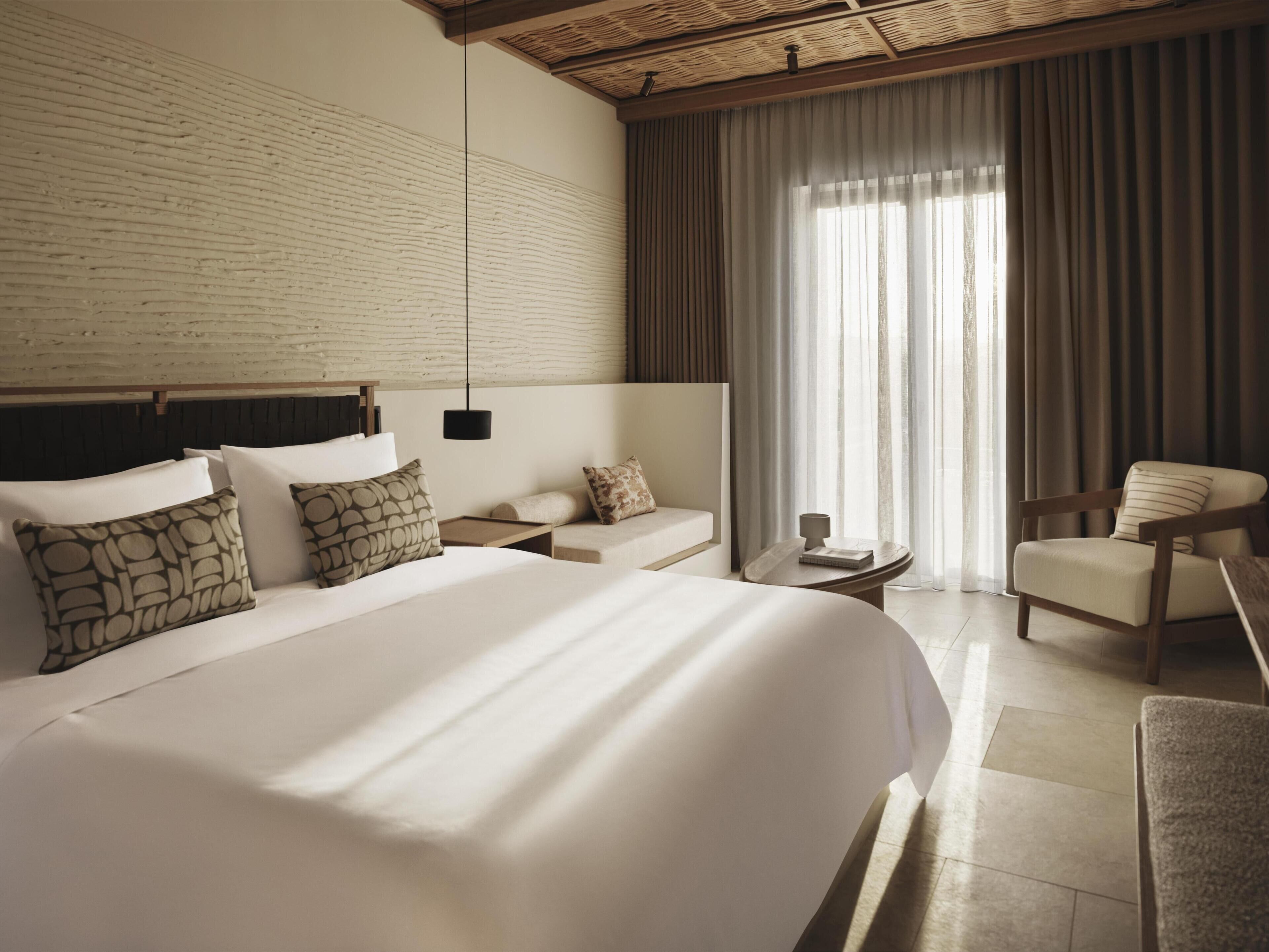 Una habitación de hotel moderna con una cama grande, un cabecero de madera, una mesita, una silla y un ventanal con cortinas.